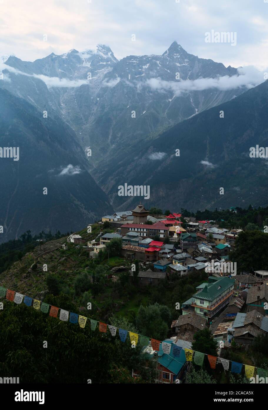 Village flanked by the high peaks of  Himalayas on a fine summer morning with Buddhist prayer flags. Kalpa,  Himachal Pradesh, India. Stock Photo