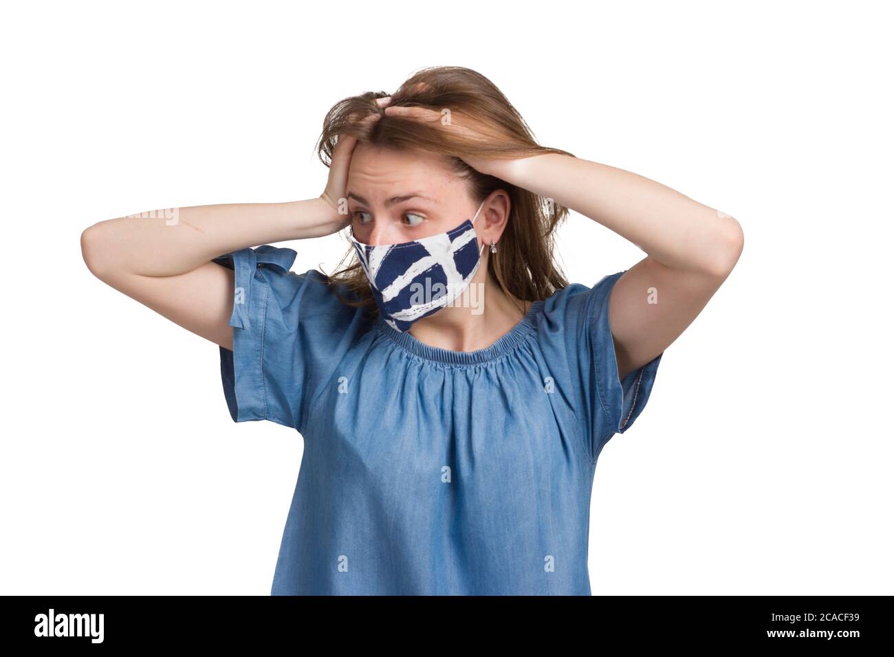 A young girl with long hair in a blue dress. Isolated on a white background. in a medical mask. holding her hands over her head, scared or panicking Stock Photo