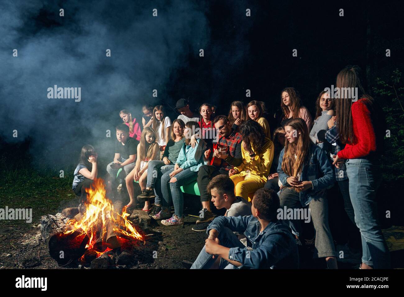 Side view of happy kids sitting on wooden bench and singing near camp fire in warm summer night. Scoutmasters playing guitar and having fun with children in forest. Concept of summer camping. Stock Photo
