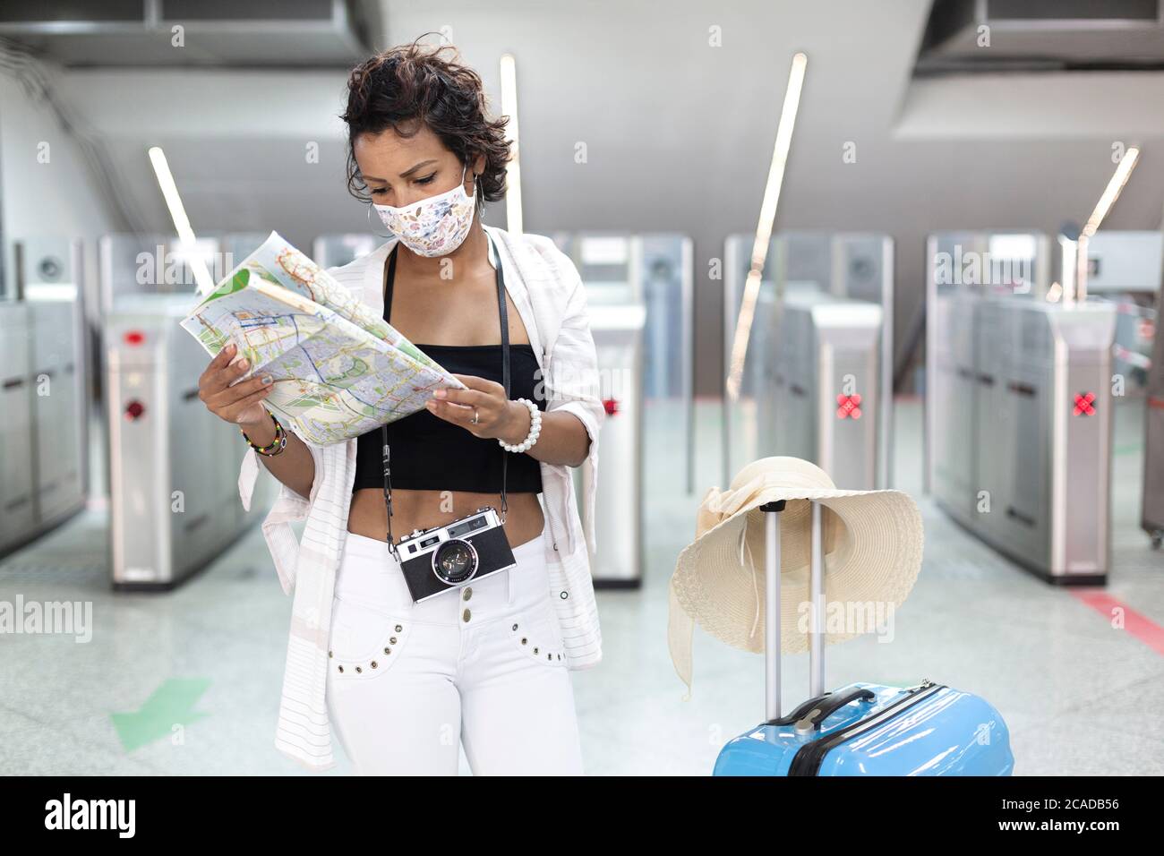 Traveling woman with a face mask looking at a tourist map. She's standing next to a travel bag. New normal travel after covid-19 pandemic concept. Stock Photo