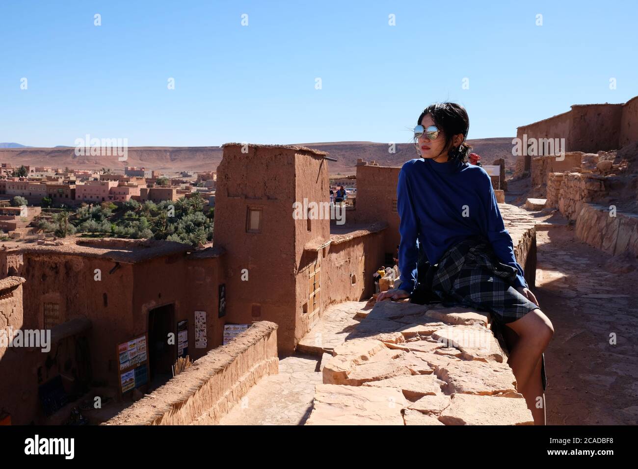 One young Asian girl sitting on old brown city wall of Ait Ben Haddou under sunshine. Famous ancient berber kasbah in Morocco Stock Photo