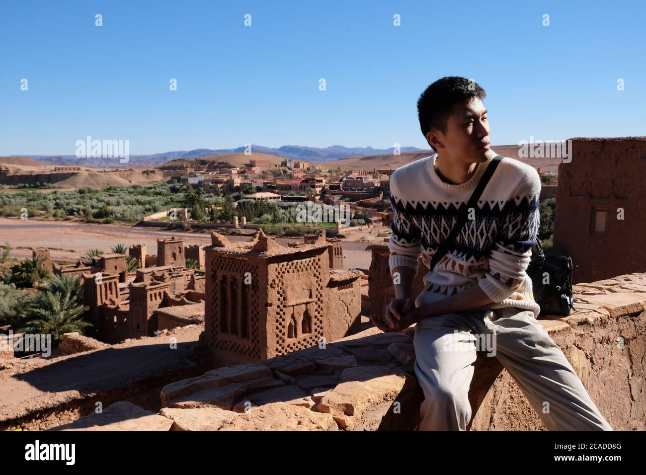 one Asian young man sitting on old brown city wall of Ait Ben Haddou under sunshine. Famous ancient berber kasbah in Morocco Stock Photo