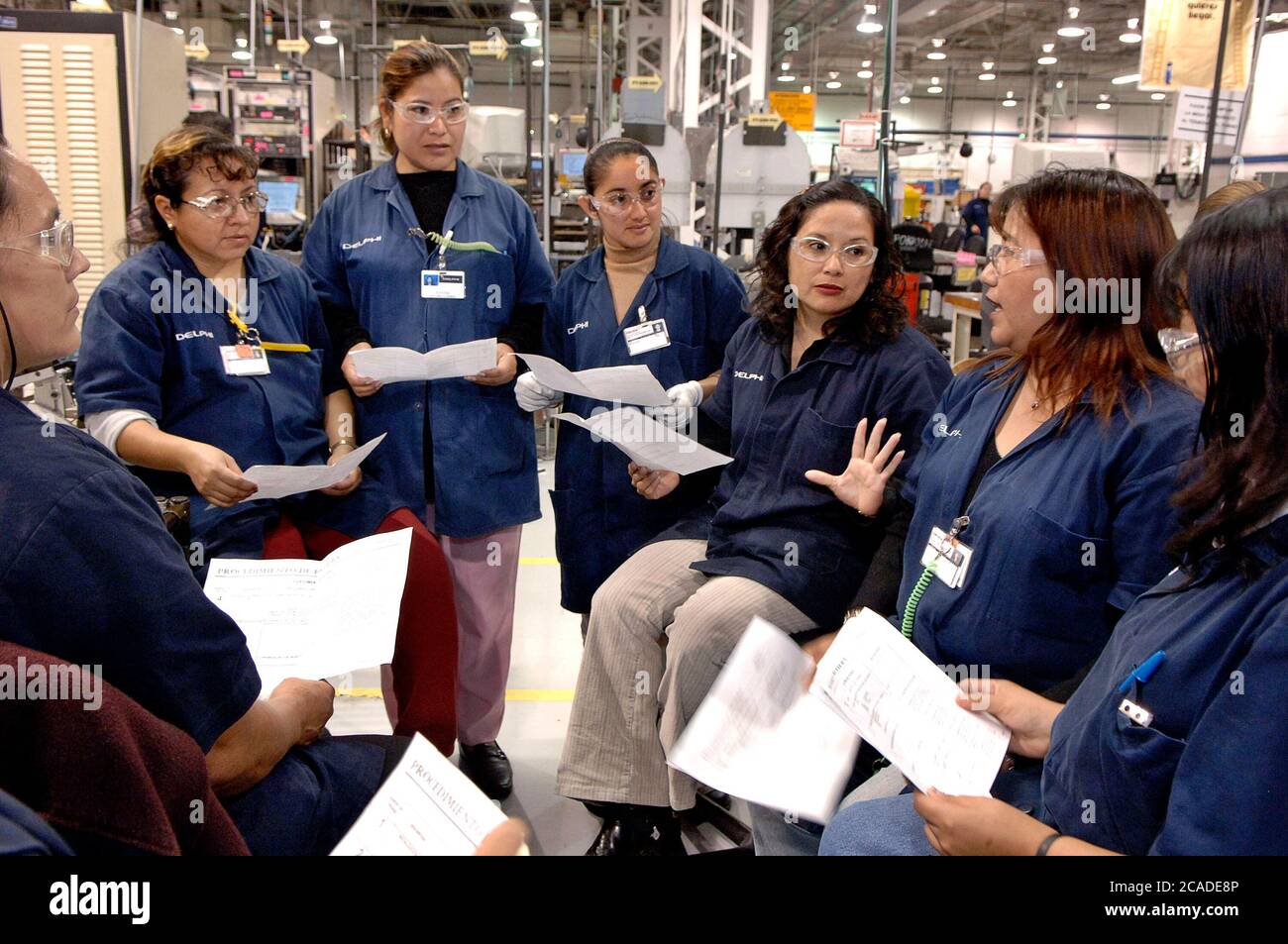 Matamoros, Mexico, April 2006: Female employees wearing lab coats and safety glasses hold a small shift meeting to discuss the day's production on the manufacturing floor at Delphi Delco Electronics de Mexico. The maquiladora plant, just across the United States border from Brownsville, Texas, makes radios and other parts for General Motors cars. Delphi has about 11,000 Mexican workers in seven factories near Matamoros.  ©Bob Daemmrich Stock Photo