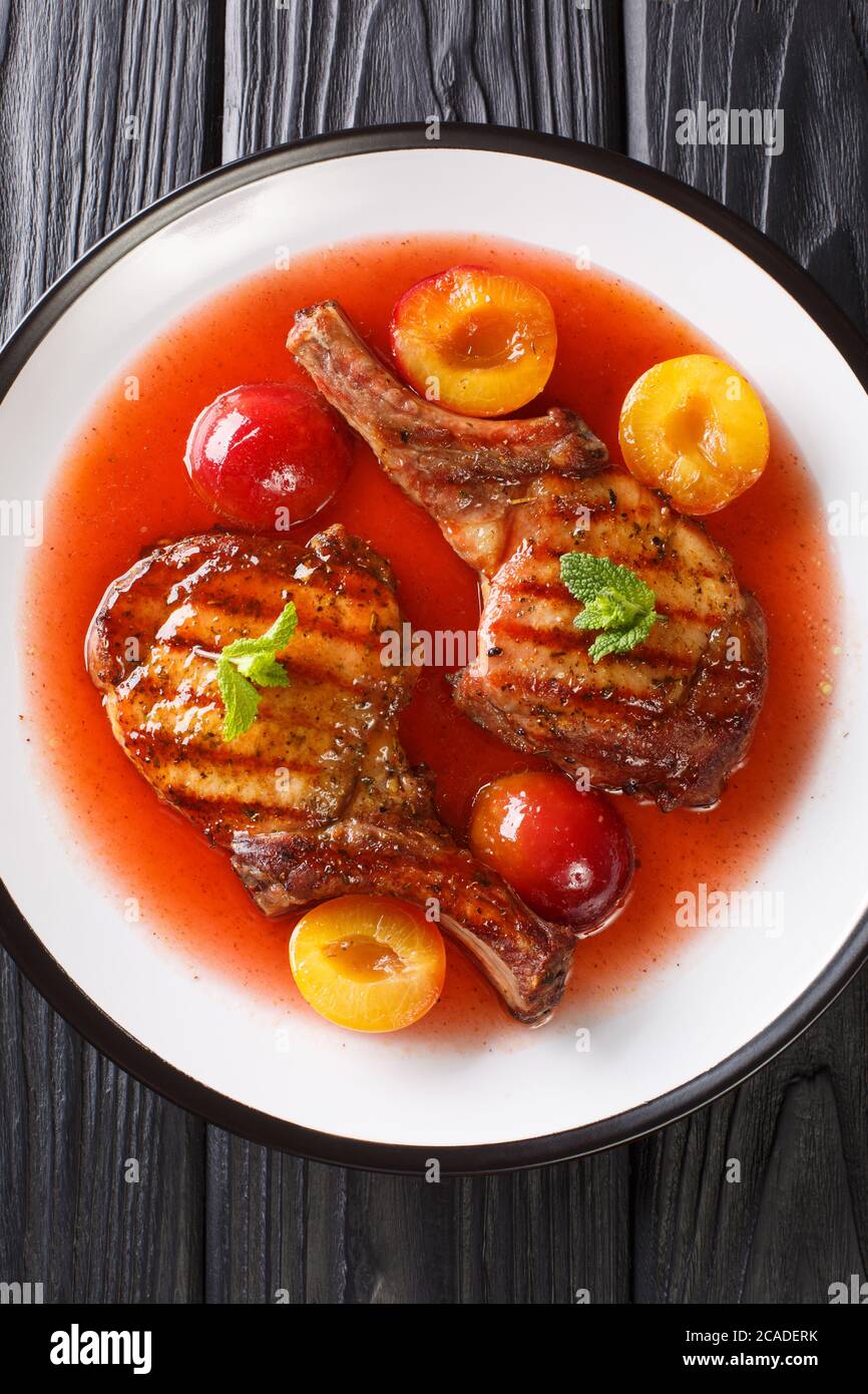 grilled pork chop with ripe plums and sauce close-up in a plate on the table. Vertical top view from above Stock Photo