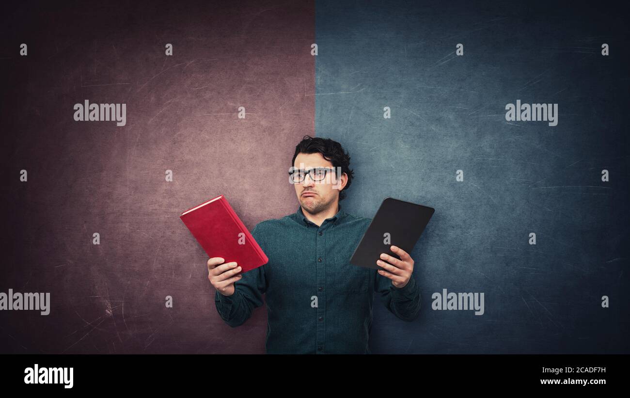 Pensive male student chooses between a traditional book and electronic PC tablet over a split wall with red and blue sides. Future education concept. Stock Photo