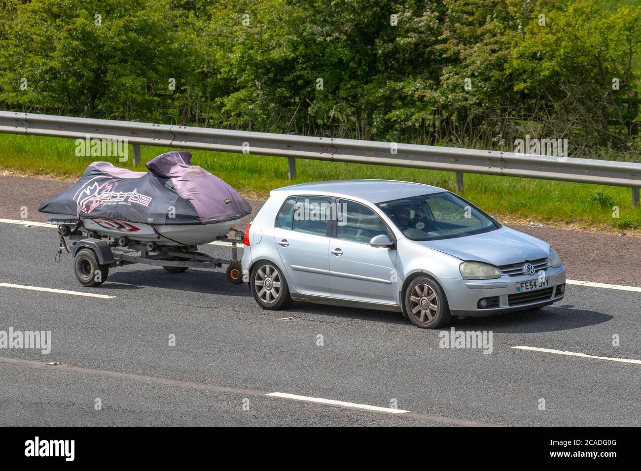 2004 silver VW Volkswagen Golf GT TDI towing covered jetskis on trailer; Vehicular traffic moving vehicles, cars driving vehicle on UK roads, motors, motoring on the M6 motorway highway network. Stock Photo