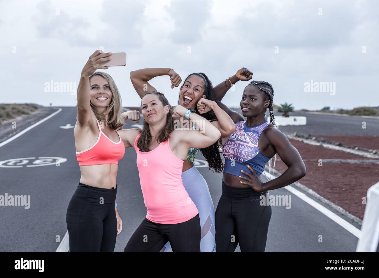 Group of positive diverse women in sportswear demonstrating biceps and smiling while taking selfie during outdoor running training Stock Photo