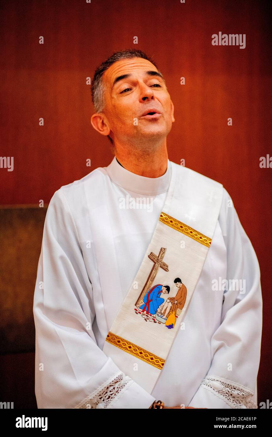 In the back of a Southern California Catholic church, a deacon in a white robe sings a hymn along with parishioners during a mass. Stock Photo