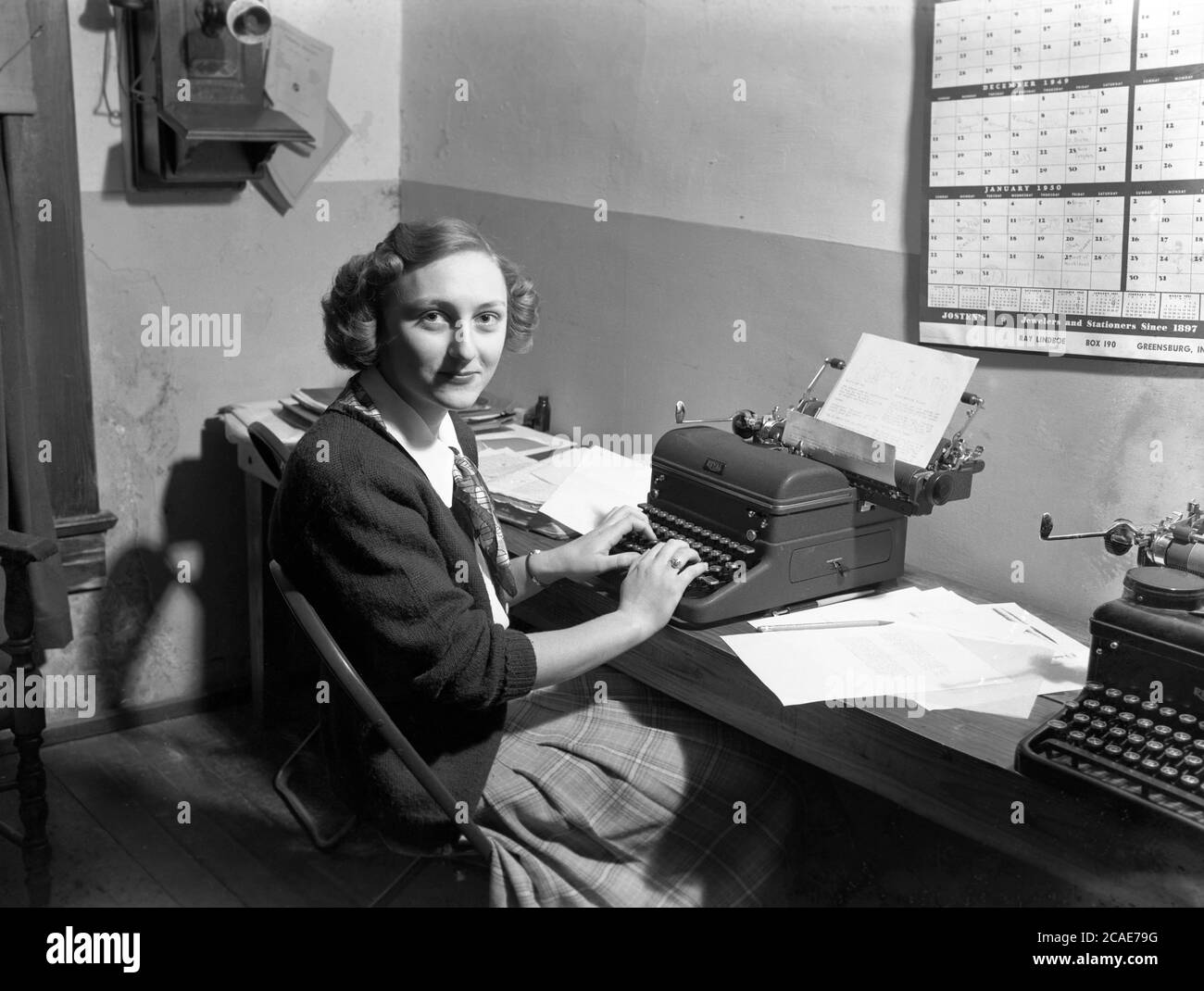 1949, historical, in a small office, a young lady sitting at a narrow wooden bench against the wall, typing a letter on a 'Royal ' typewriter of the era, USA. An internal speaker of the era is can be seen on the wall. Stock Photo