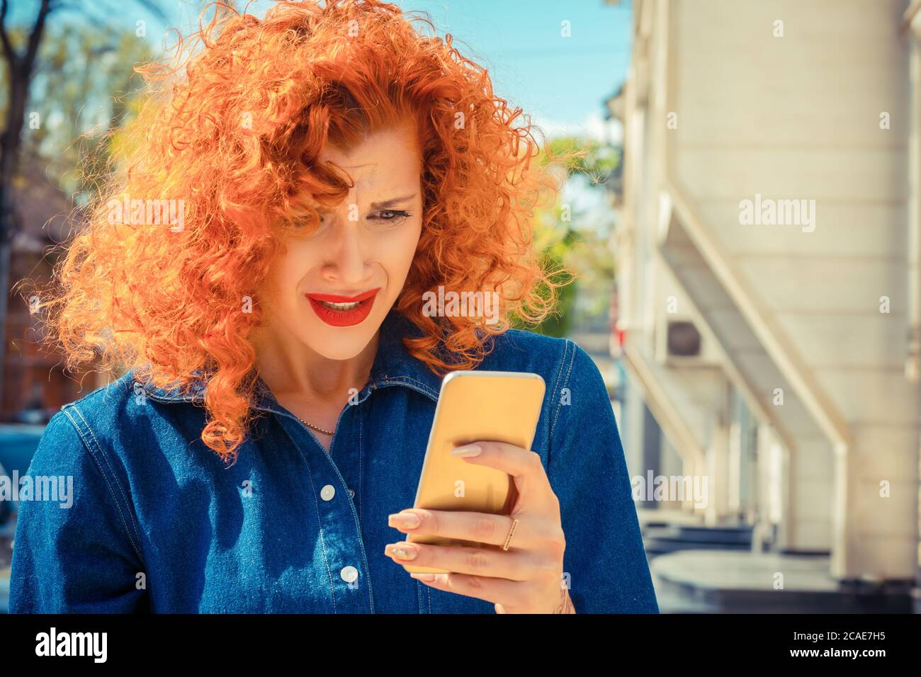 What. Unhappy frustrated angry woman with red curly hair looking to mobile phone, reading sms or something, standing outside city on background. Negat Stock Photo