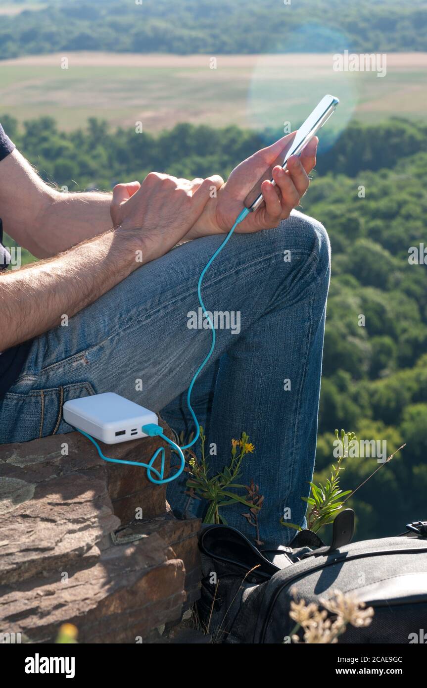 Man on a hike uses smartphone while charging from the power bank on the rock at dawn. Healthy lifestyle and communication Stock Photo