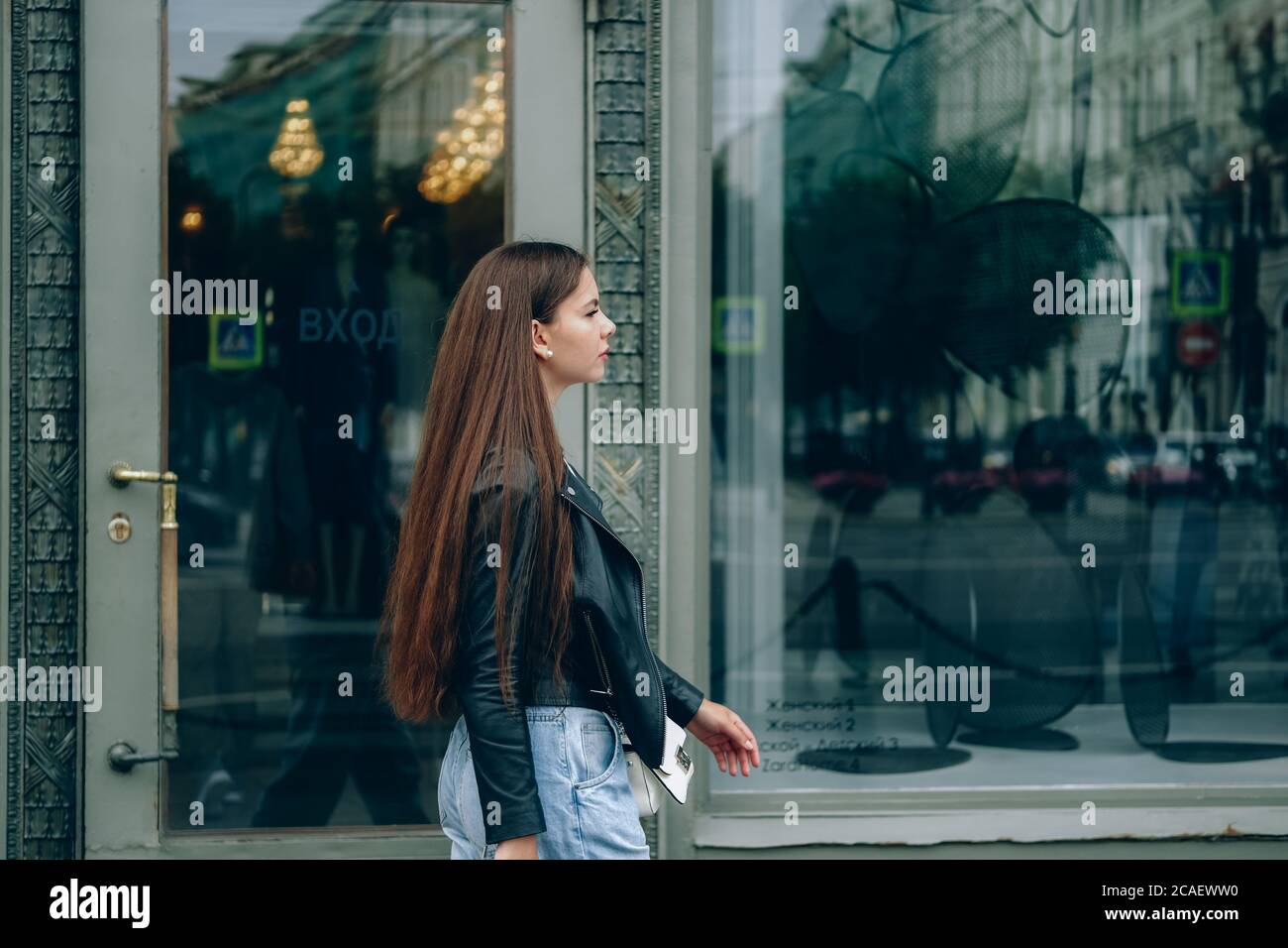 Young woman with long hair walking along the street of a big city, reflected in big shop windows. Saint Petersburg, Russia. Stock Photo
