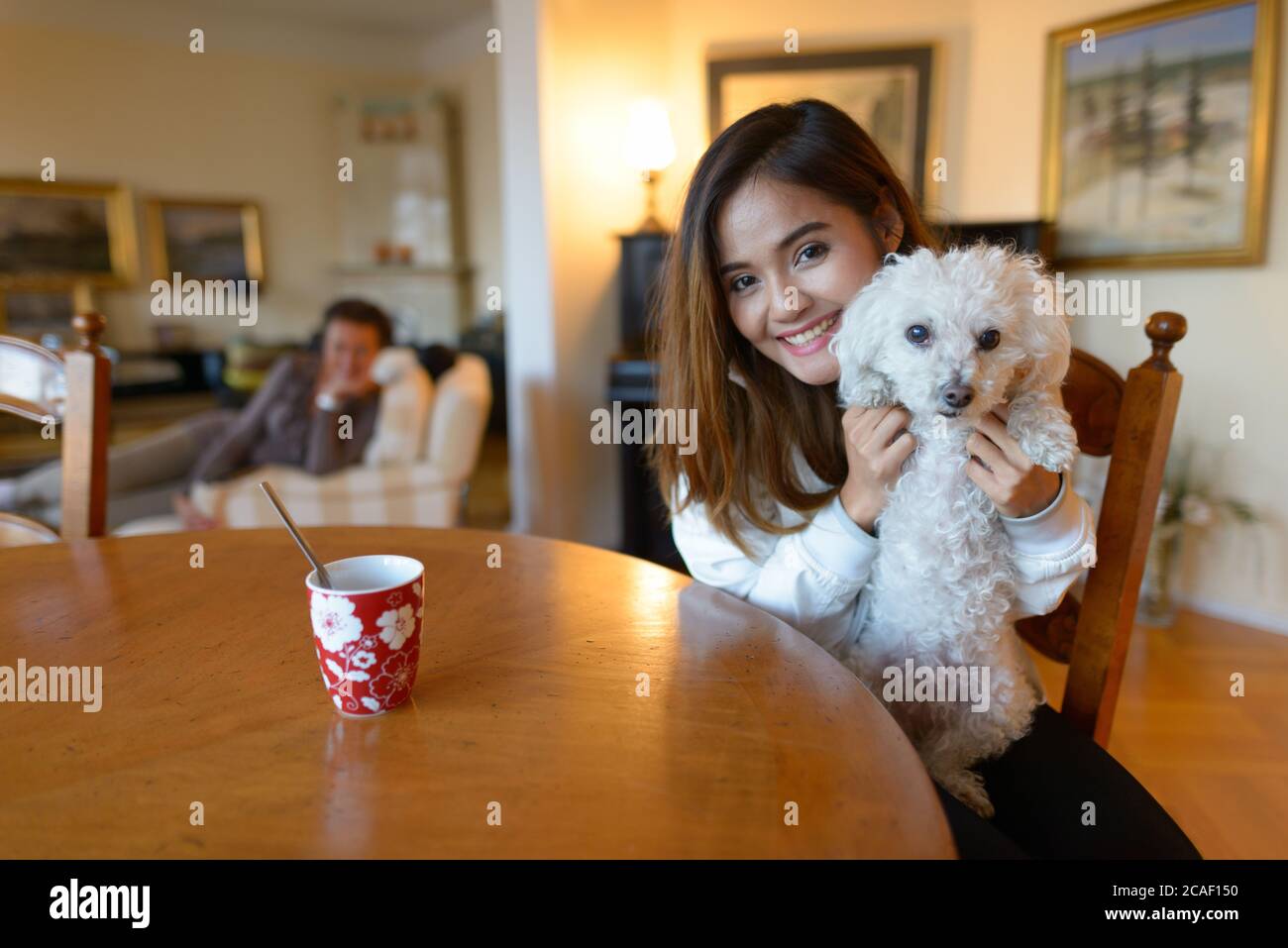 Young happy Asian woman smiling while holding cute dog inside comfortable home Stock Photo