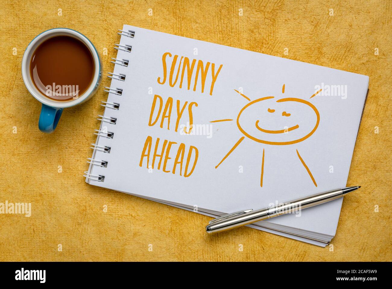 sunny days ahead inspirational note and sketch - handwriting in spiral art sketchbook against yellow textured bark paper with a cup of coffee, optimis Stock Photo