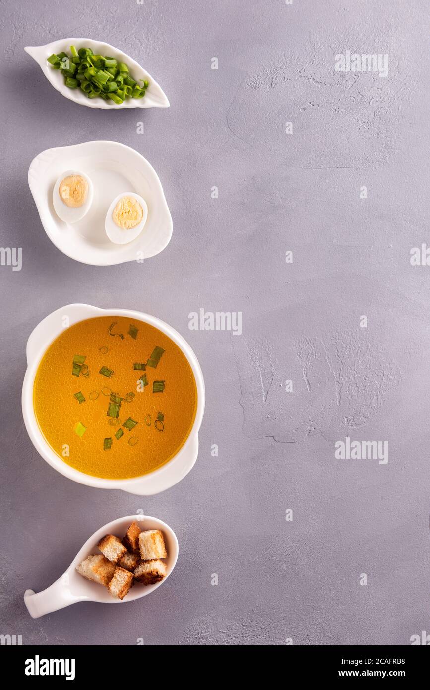 Vertical background with row of white crockery with chicken bouillon, chopped green onion, croutons and quail egg cutted on halfs on textured gray sur Stock Photo