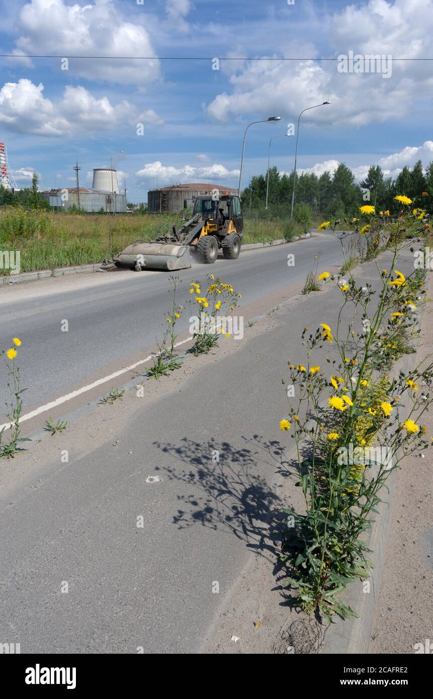 Tractor with a bucket removes garbage and a sidewalk with yellow flowers in the foreground in the background a thermal power plant Stock Photo