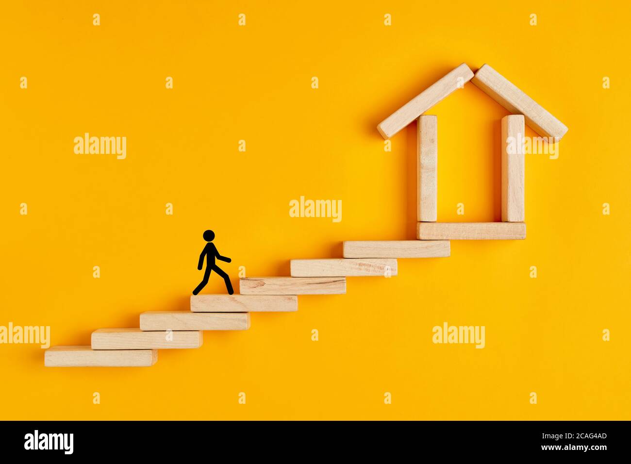 Male stickman climbing the ladders towards home formed by wooden blocks on yellow background. Stock Photo