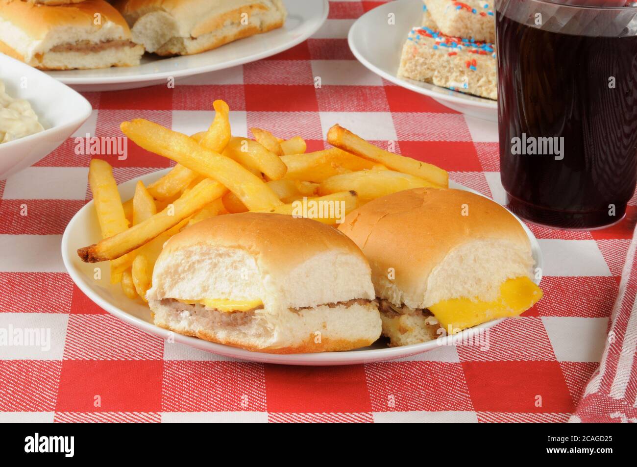 Mini cheeseburgers and fries on a picnic table loaded with food Stock Photo
