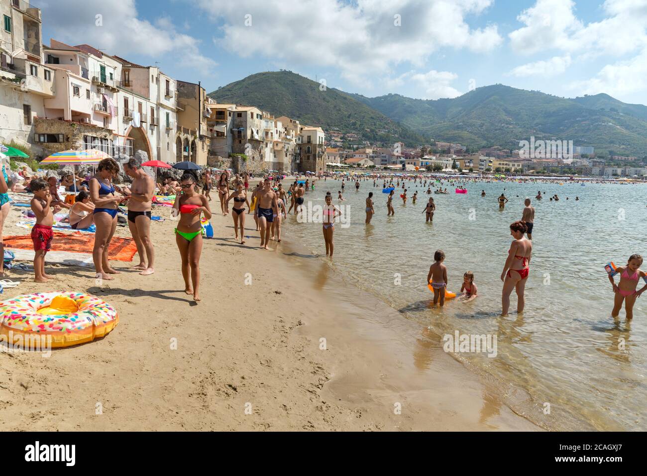 11.08.2018, Cefalu, Sicily, Italy - Tourists and locals relax on the city beach under umbrellas and swim in the Mediterranean Sea. In the background t Stock Photo
