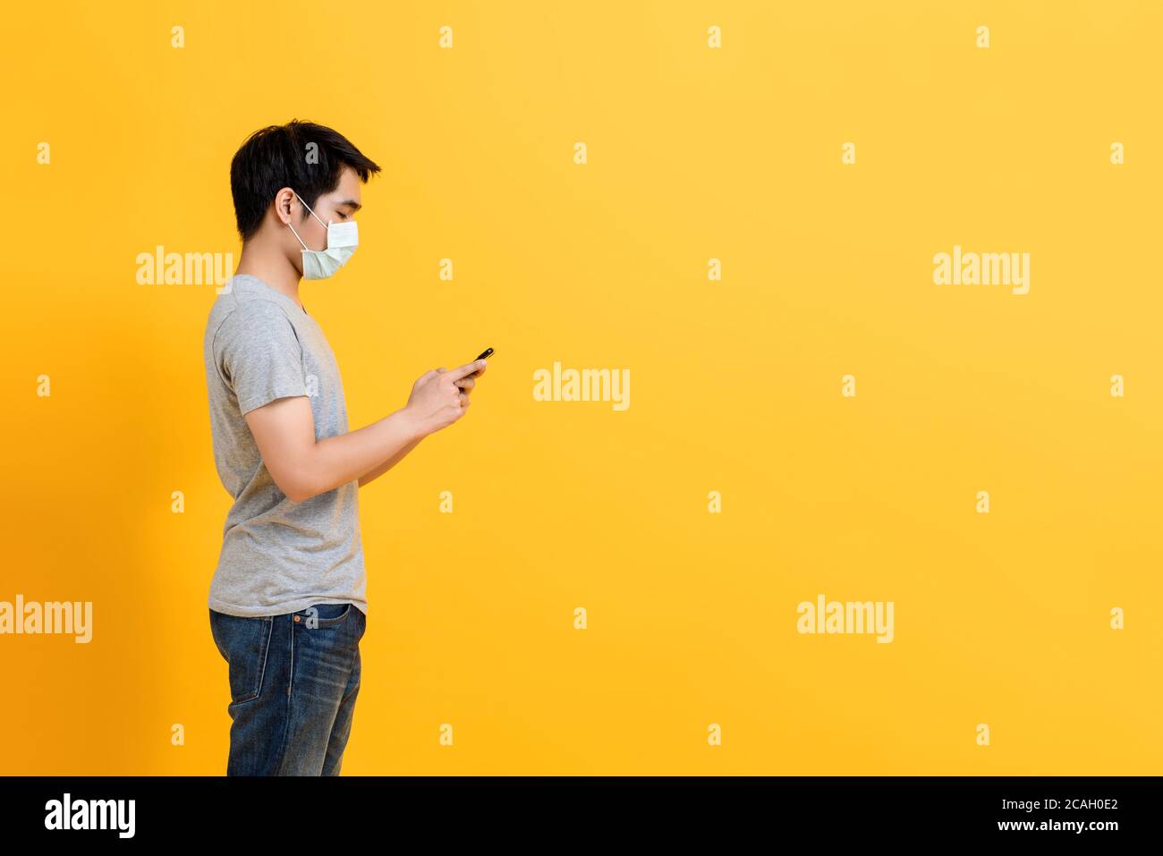 Young Asian man wearing medical face mask standing and using mobile phone isolated on yellow studio background with copy space Stock Photo