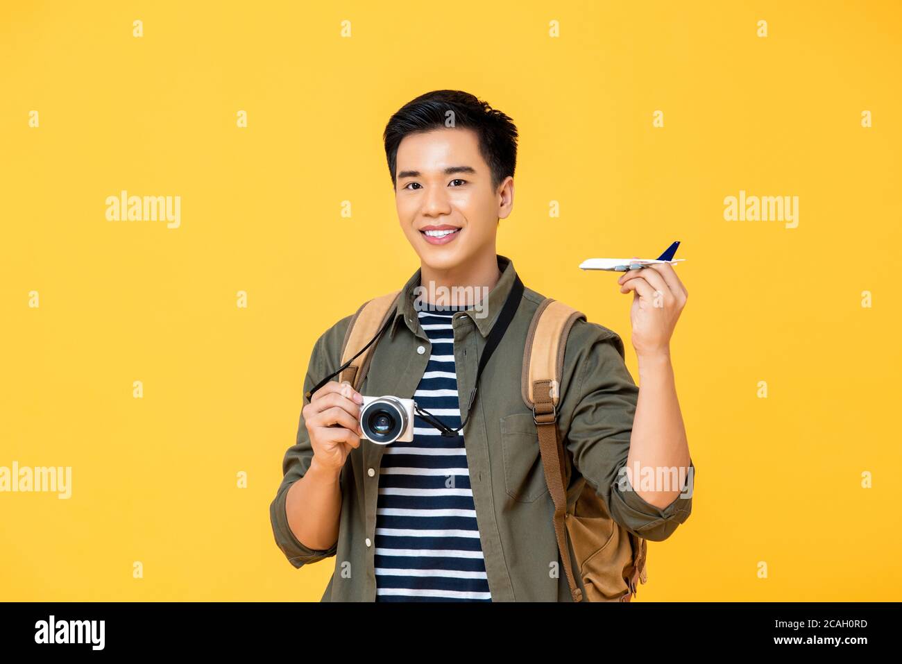 Handsome smiling Asian tourist man holding plane model and camera isolated on yellow studio background Stock Photo