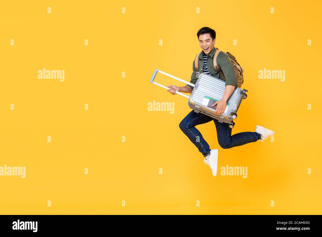 Young excited Asian tourist man with baggage jumping in mid-air ready to travel isolated on yellow background Stock Photo