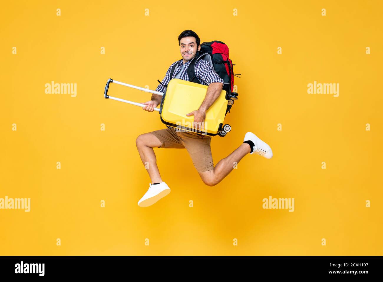 Young excited Caucasian male tourist with baggage jumping in mid-air ready to travel isolated on colorful studio yellow background Stock Photo