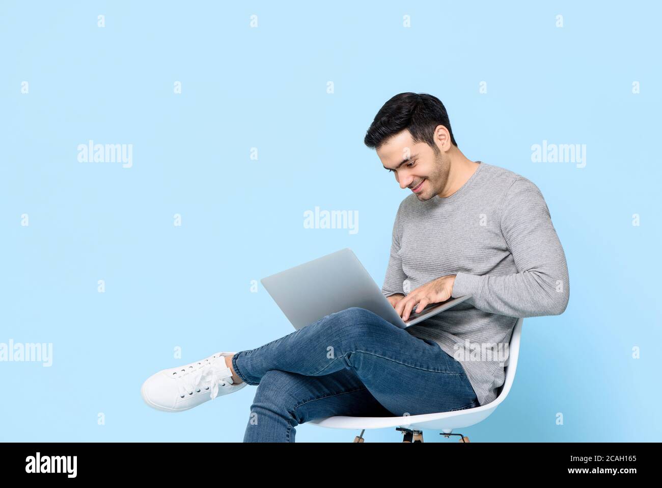 Work from home concept portrait of happy young handsome Caucasian man sitting and using laptop in isolated studio blue background Stock Photo