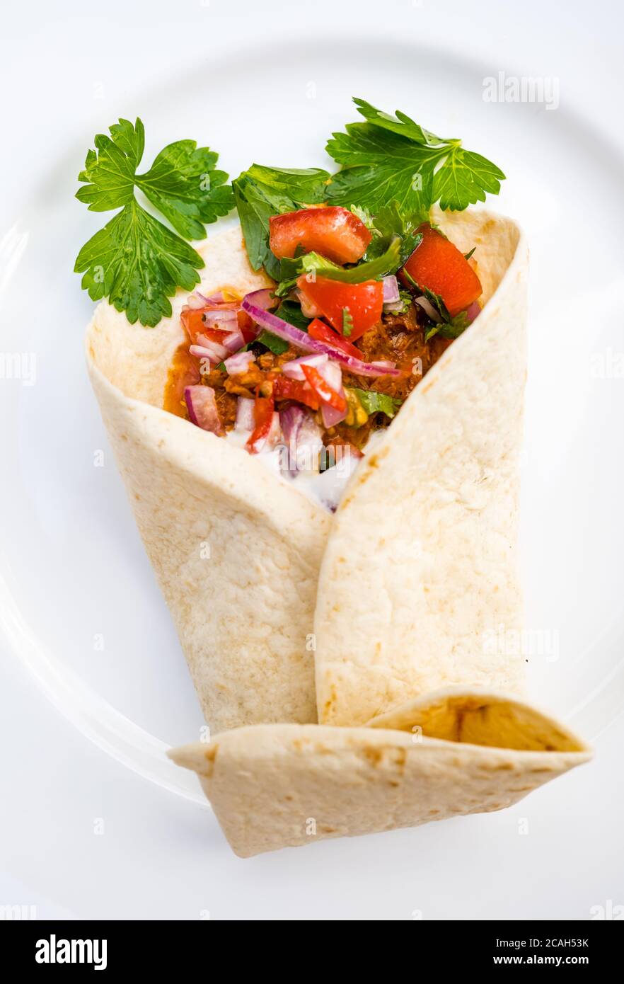 Close up of Mexican tortilla wrap filled with pulled pork, tomato salsa and sour cream with coriander on white dinner plate Stock Photo