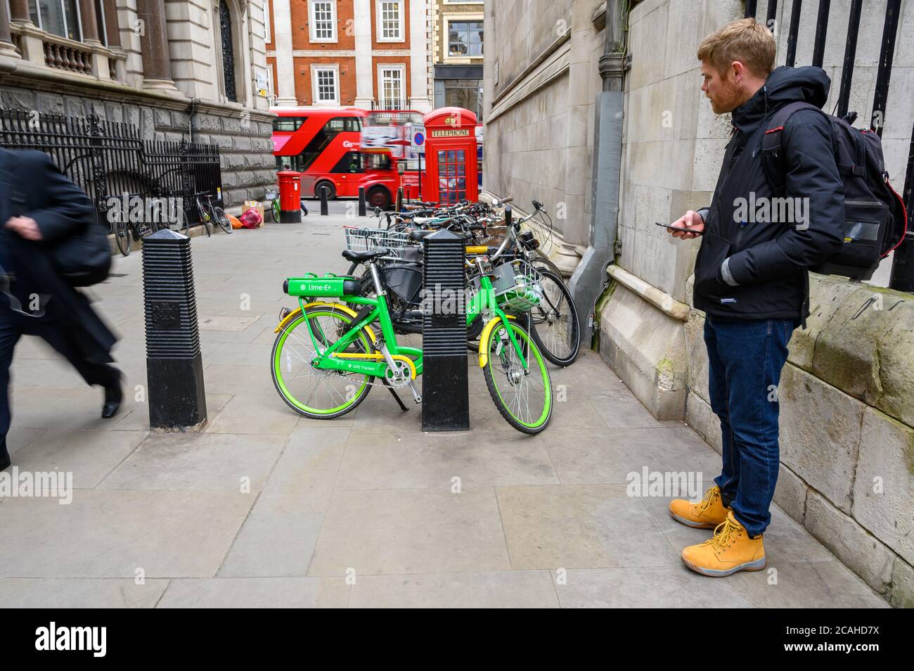 LONDON - FEBRUARY 04, 2020: A man operating a smartphone and looking at a Lime electric assist rental bike. Red London Double Decker Buses passing in Stock Photo