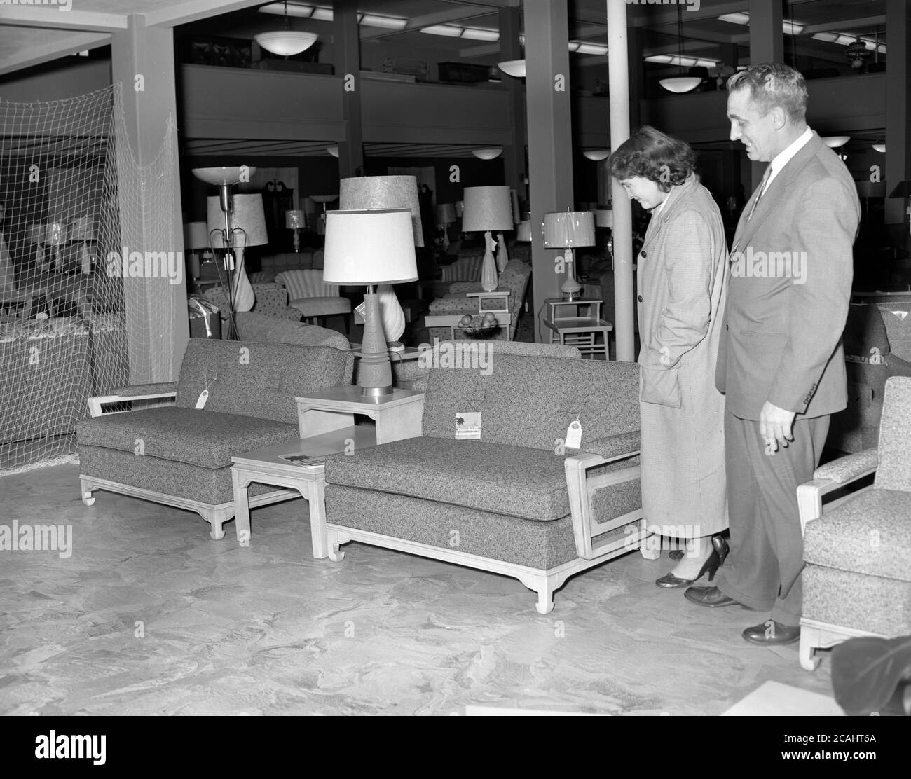 circa 1940s, historical, a lady in a large furniture and home interiors store looking at a pair of moern, small two-seater sofas, with suited male customer representative in attendance, USA. Stock Photo