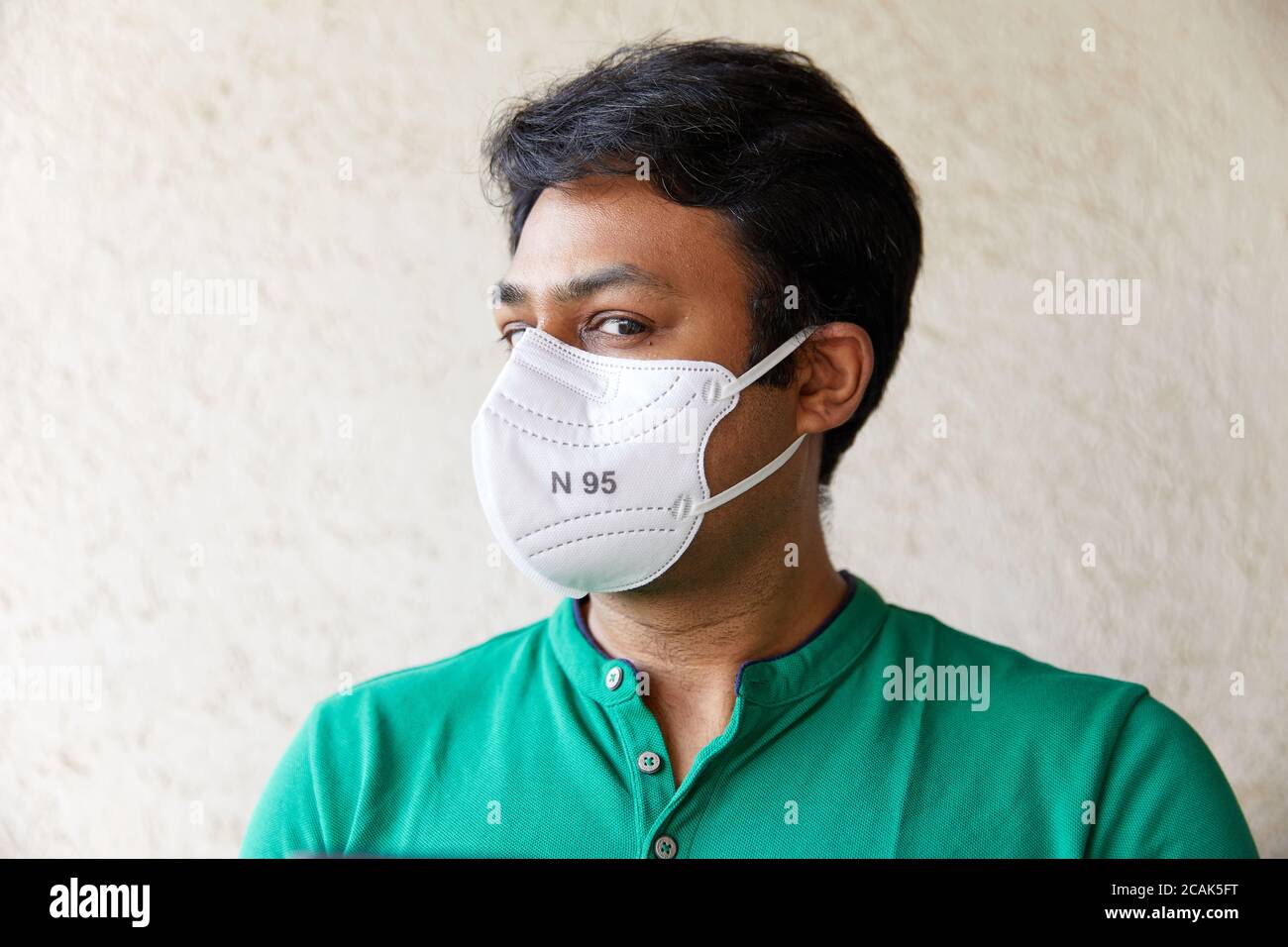 Adult Indian Male wearing a White N95 Respirator in Green T-Shirt looking into the camera with head faced 45 degrees with textured background Stock Photo