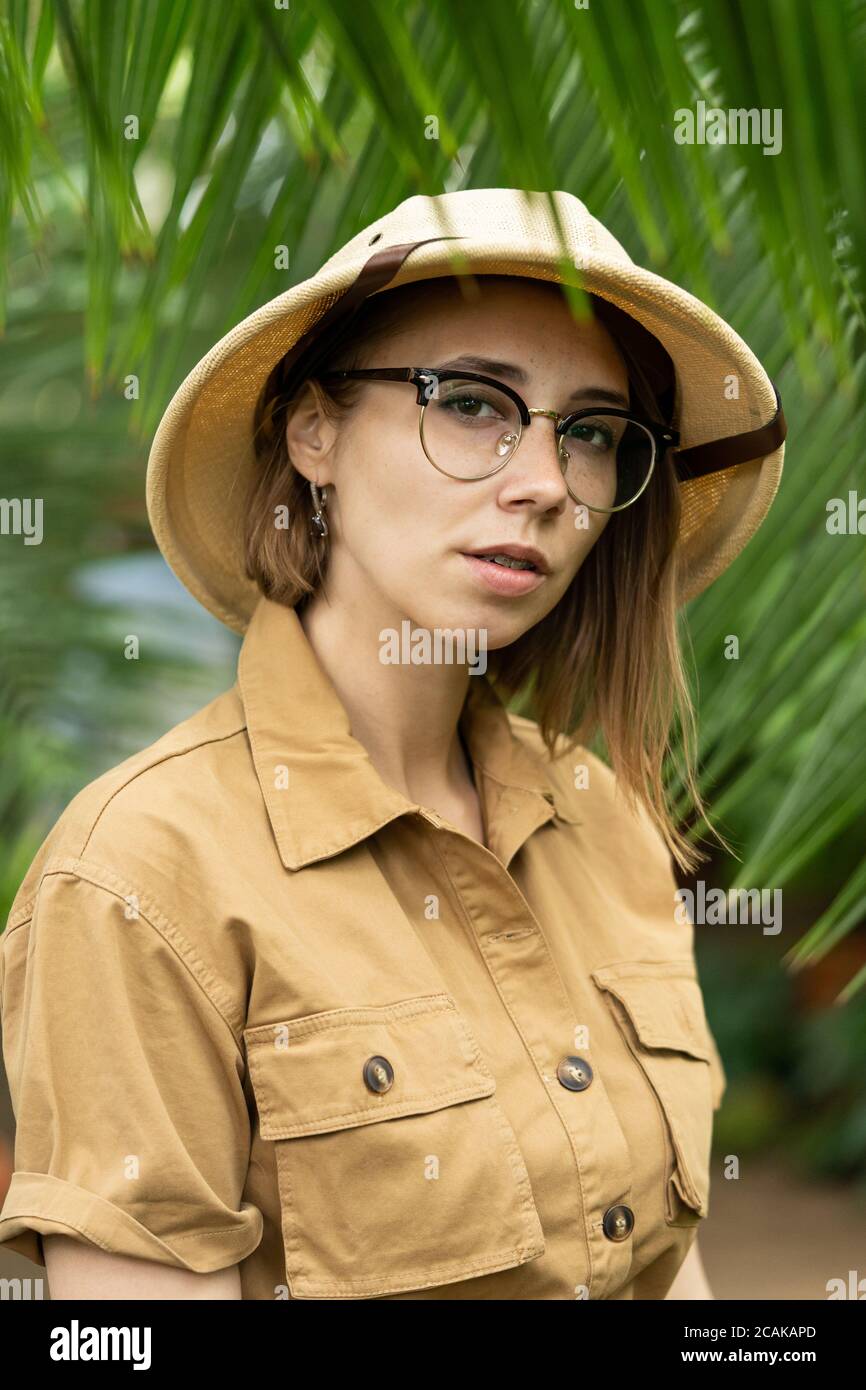 Woman botanist dressed in safari style in greenhouse. Naturalist in khaki clothes, leaver gloves in rainforest surrounded by palms, looking at camera. Stock Photo