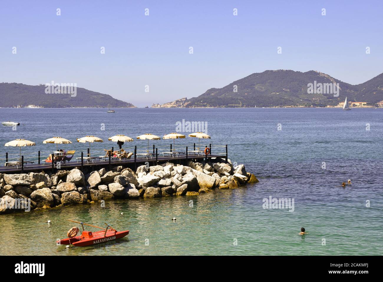 View of the Gulf of the Poets with people sunbathing on the rocky shore, the promontory of Porto Venere and the Palmaria Island, Lerici, Italy Stock Photo