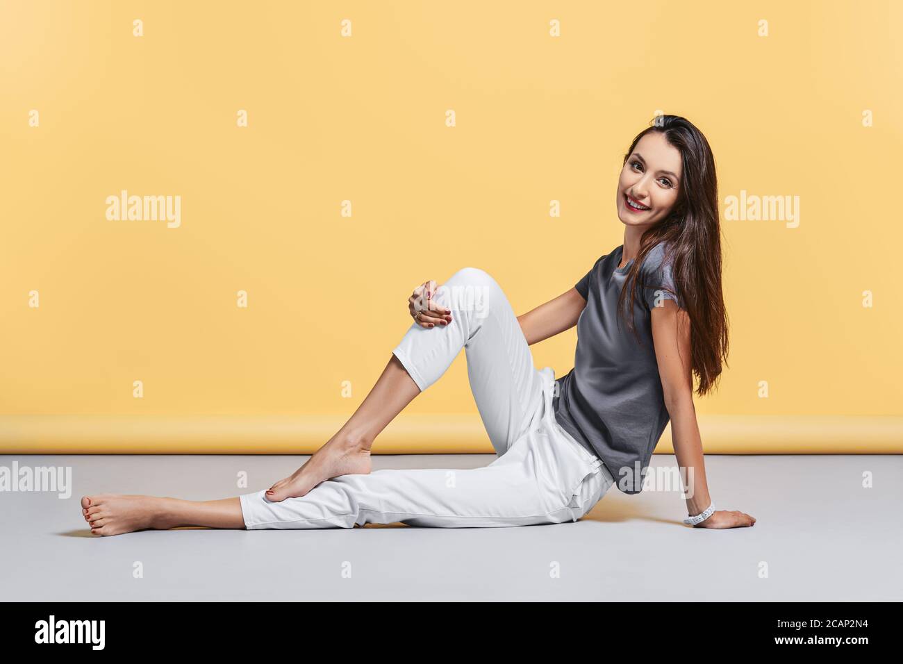Happy barefoot young woman in casual trousers and shirt sits on the floor in studio on yellow background Stock Photo