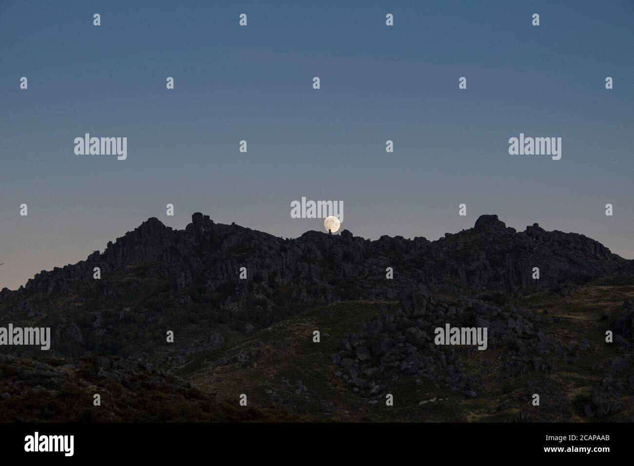 Almost full moon slowly rises behind stones on mountain peaks in a twilight moment, far away in the distance Stock Photo