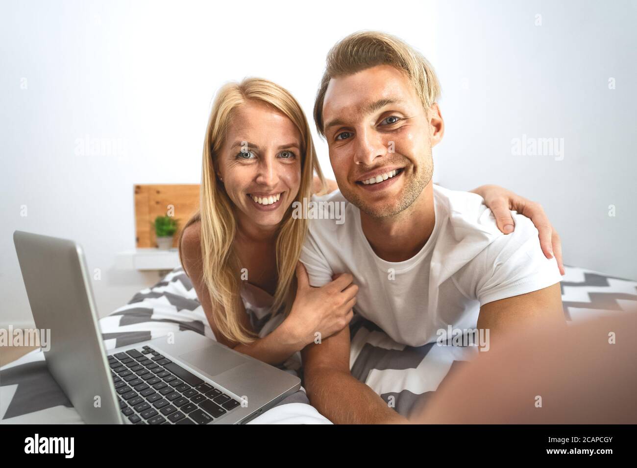 Young couple taking selfie in bed - Happy lovers having fun taking self photos lying on bed while using laptop in bedroom Stock Photo