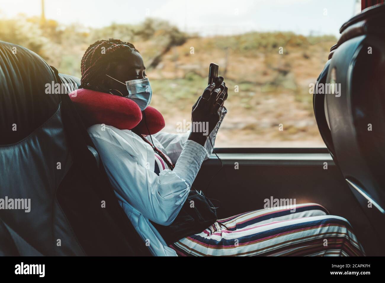 Side view of a black woman tourist using a smartphone on a seat of a modern regular bus near the window, she has a red air cushion on her neck Stock Photo