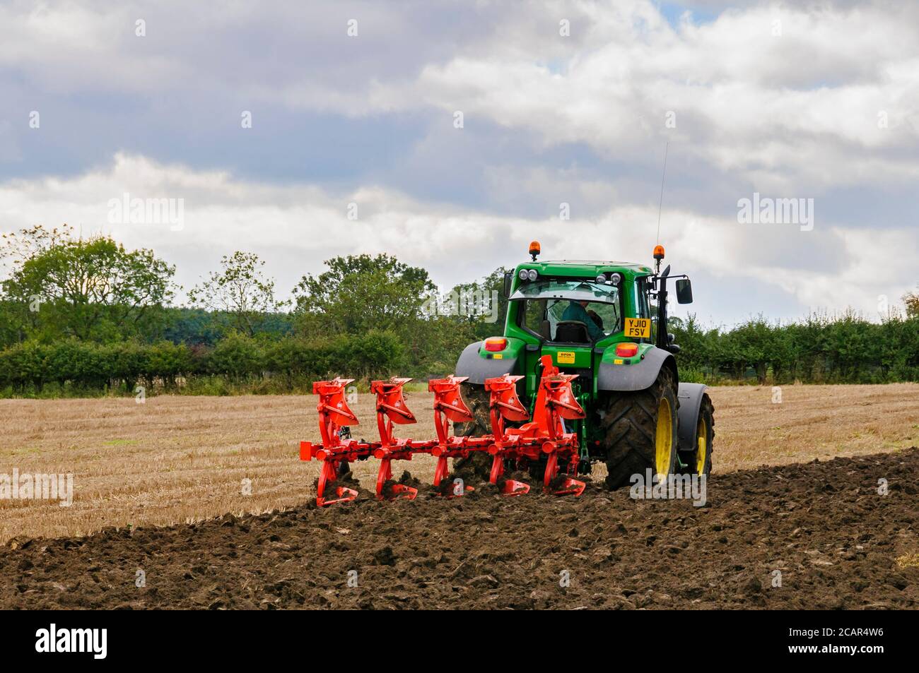 Green farm tractor working in field, pulling 5-furrow reversible plough,  stubble ploughing & turning soil (farmer driving) - North Yorkshire, GB. Stock Photo