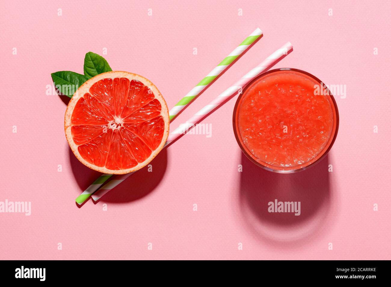 Glass of freshly squeezed grapefruit juice with pulp, half grapefruit and two drinking straws cast shadow on a pink background. Hard light shot. Stock Photo