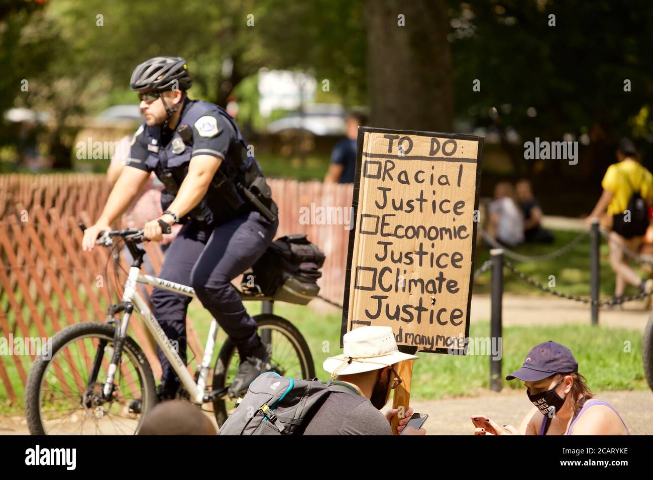 Washington, DC, USA. 8th Aug, 2020. Pictured: Protester holds a sign listing the social and legal changes needed as one of the 10  Metropolitan Police (DC police) rides by on a bike. Credit: Allison C Bailey/Alamy. Credit: Allison Bailey/Alamy Live News Stock Photo