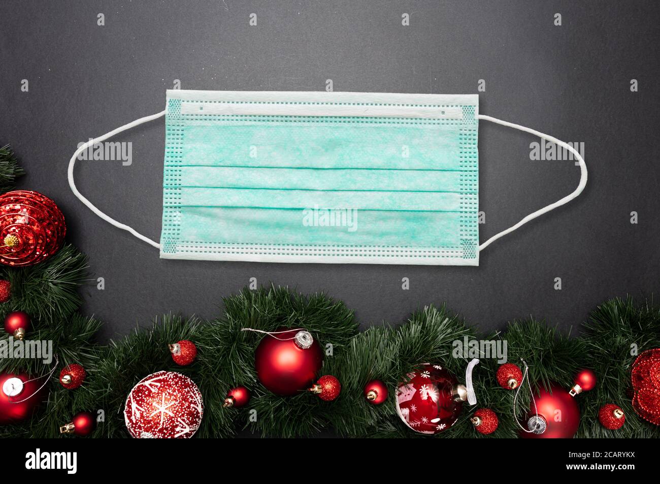 Coronavirus and Christmas 2020 concept. Face protective mask and xmas festive garland with red baubles against gray black background. COVID19 spread p Stock Photo