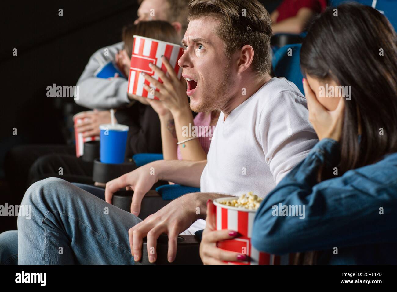 Good lord! Young man looking extremely shocked watching scary movies with the group of his friends at the local movie theatre Stock Photo
