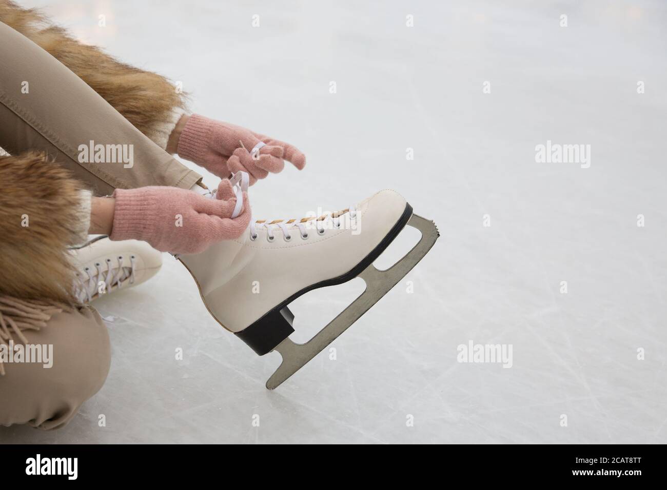 Closeup of female in fur coat and pink gloves wearing white skates on ice rink in winter day.Woman tying skates.Weekends activities outdoor in cold we Stock Photo