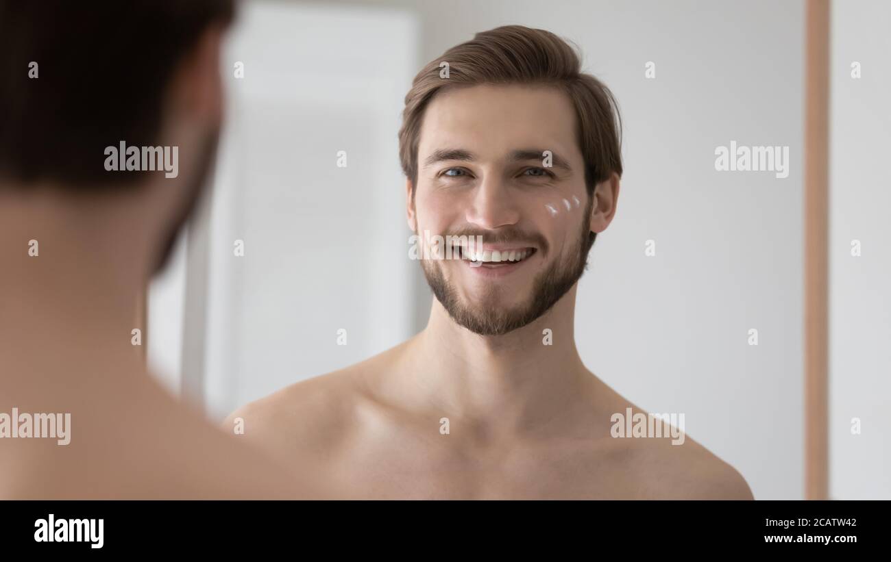 Head shot mirror reflection smiling young man applying moisturizing cream Stock Photo