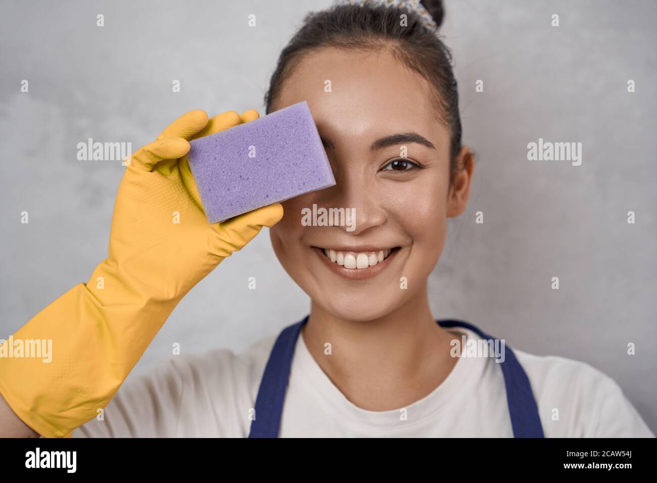 Close up portrait of funny cleaning lady in yellow rubber gloves covering one eye with kitchen sponge, looking at camera and smiling, standing against grey wall. Housekeeping, cleaning services Stock Photo