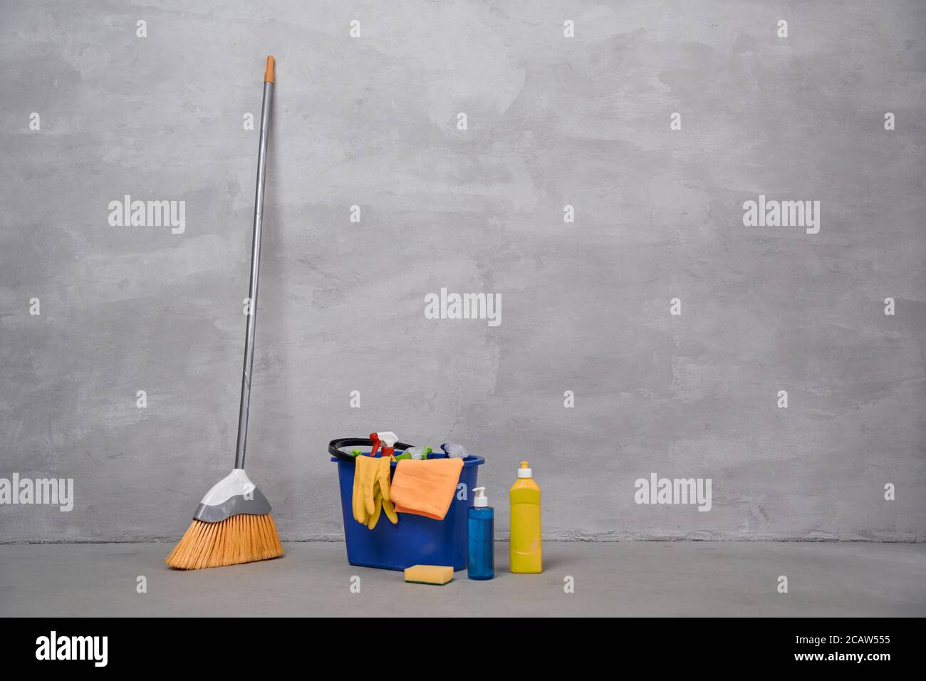 Cleaning equipment. Broom and plastic bucket or basket with cleaning products, bottles with detergents standing on the floor against grey wall. Housework, cleaning, housekeeping concept Stock Photo