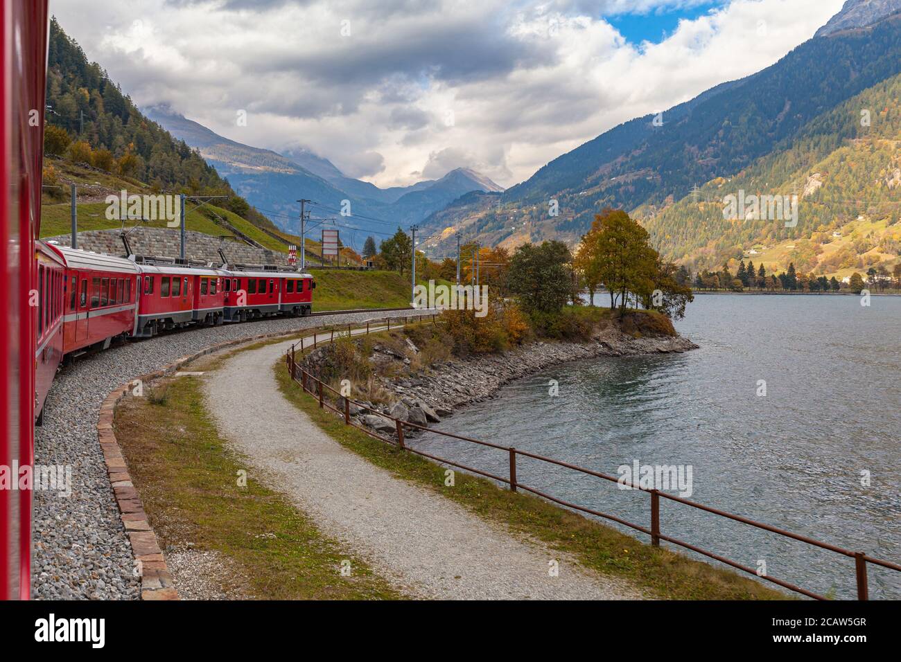 Beautiful view of  red  Rhaetian train running on the lake side of Lago di Poschiavo in autumn with blue sky cloud, on sightseeing railway line Bernin Stock Photo