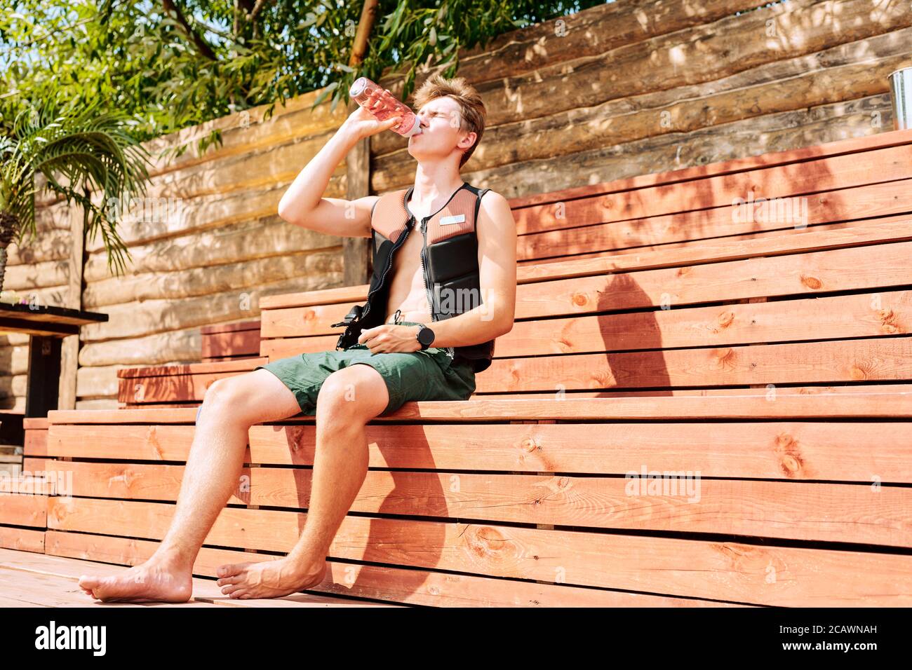 Young athlete in shorts and safety jacket sitting on wooden bench on sunny day Stock Photo