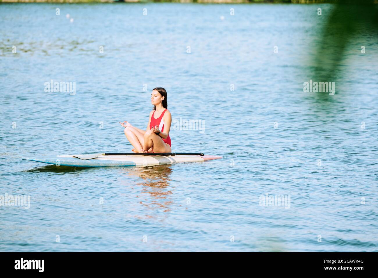 Young serene female in red swimsuit sitting on surfboard in pose of lotus Stock Photo